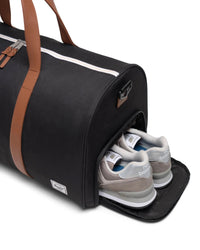 Black duffel bag with brown straps containing a pair of gray sneakers on a white background